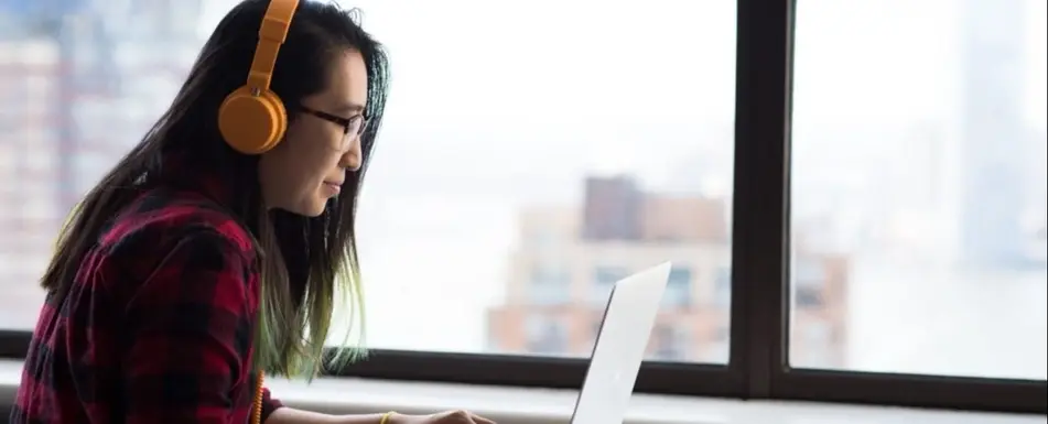 A woman looking at her laptop. She is wearing orange headphones.