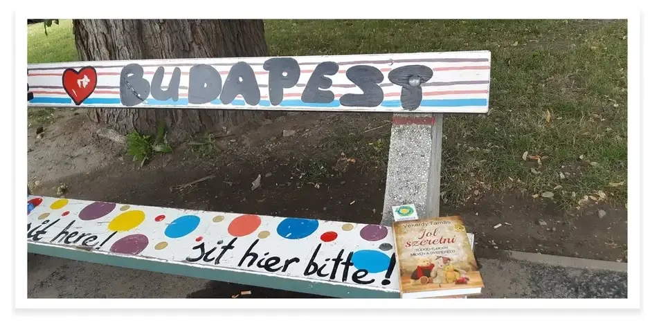 A photograph of Magdolna R.'s Book on a Bench project in Budapest. A small book is placed on the corner of a brightly-painted bench, which has the words "Budapest" painted along the back.