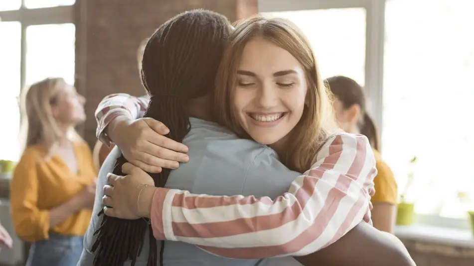 Duas pessoas felizes se abraçando em um ambiente iluminado, com outras pessoas ao fundo.