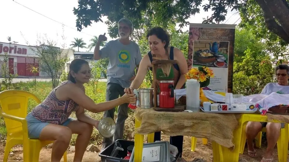 Imagem de 4 pessoas, sendo uma mulher servindo um café à outra, um homem com a camiseta idealista e outra mulher sentada na ponta da mesa.