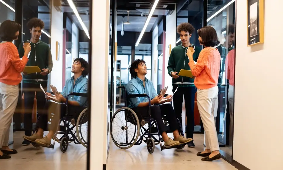 A photograph of a man in a wheelchair holding interview materials as he chats with two people in the hallway of an office.