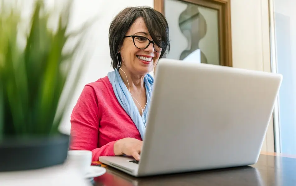 An older woman sits at a desk typing on her computer while smiling.