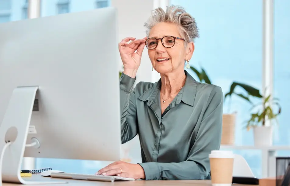 Older job seeker with short hair and glasses sits at a computer in her office.