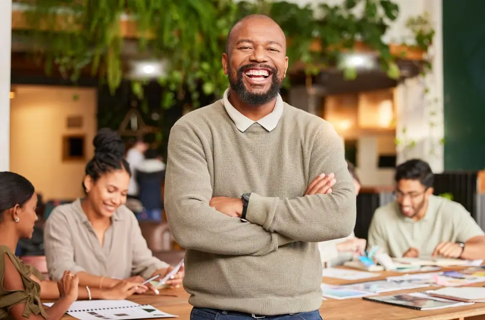 A photograph of an older Black man smiling as he stands in front of a long desk, with co-workers sitting and smiling behind him.