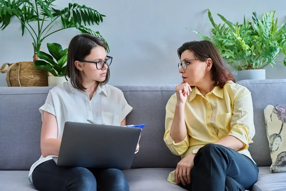 Two white women sit on a grey couch discussing salary information, with green plants in the background.