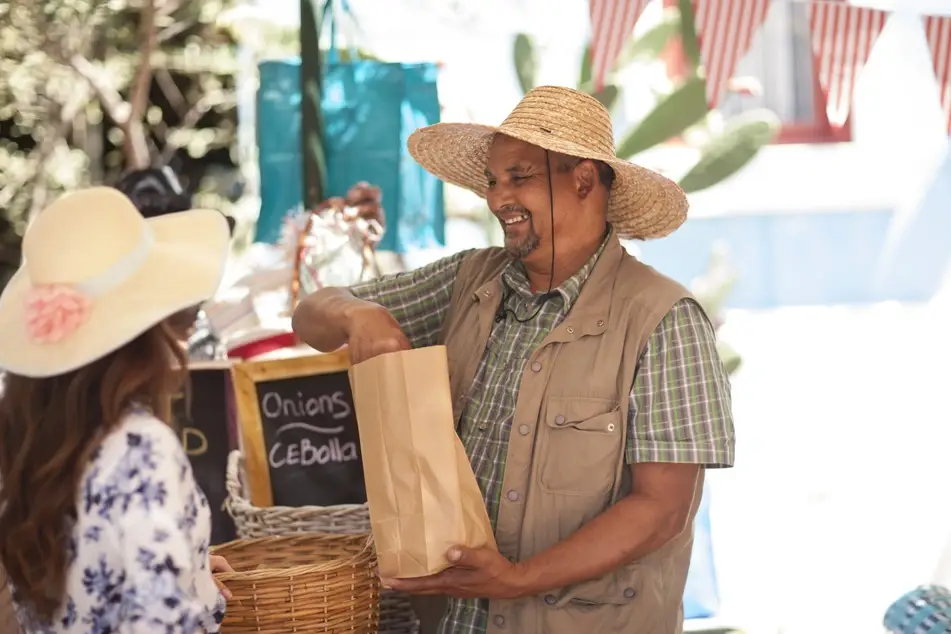 A man wearing a sun hat packages produce at the farmer's market for a customer.