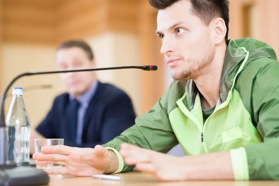 A young white man wearing a green windbreaker sits at a city council meeting to participate in his community.