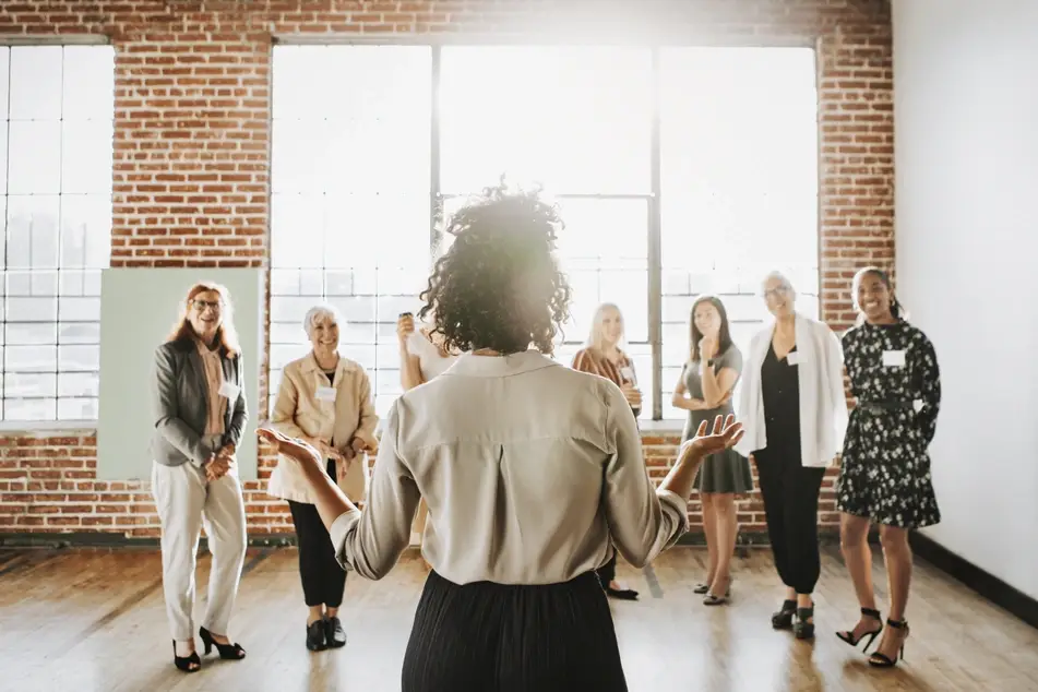 A group of multiracial and multigenerational women stand inside of a bright empty room with brick walls. They all have name tags and are smiling at a woman standing at the front of the room.