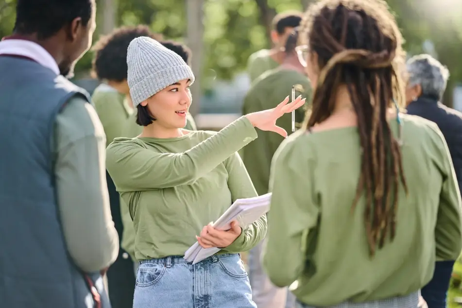 An image of a young woman with short brown hair directing a group of volunteers. They are all wearing green long-sleeve tshirts and standing around outside.