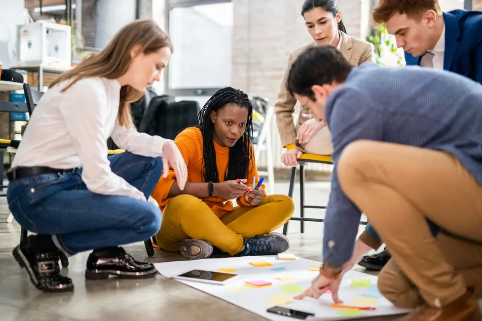 A Black woman wearing a yellow sweater and pants sits on the floor with her colleagues, planning professional development with post-it notes and a white sheet of paper.