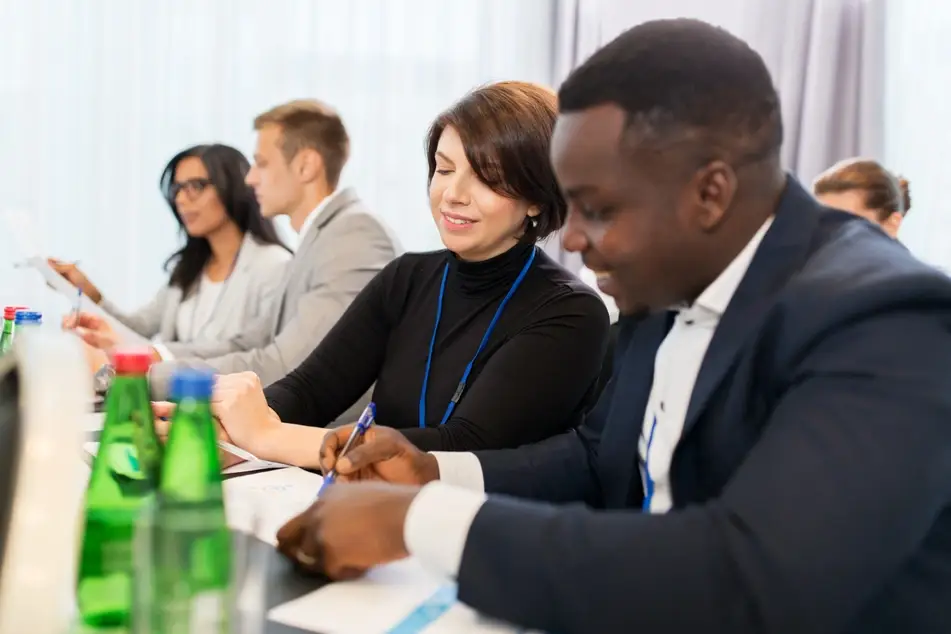 Black businessman and white businesswoman chat at a nonprofit board meeting with colleagues, green water bottles on a long table.