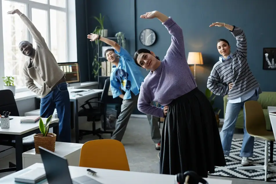 Group of co-workers stretch in office to prevent work burnout by having fun together.