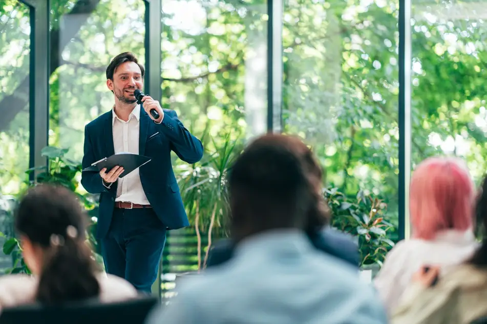 A man in a blue suit holds a microphone and speaks to an open conference room, with large windows with trees in the backgound.