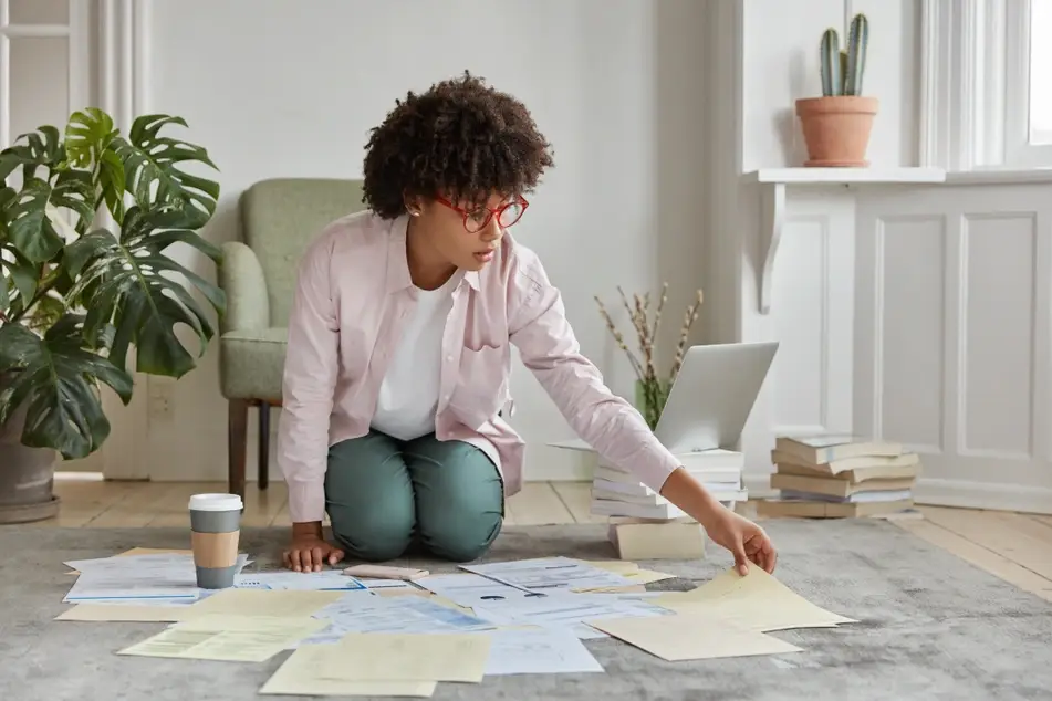 Black woman wearing glasses and button down kneels on rug surrounded by papers.