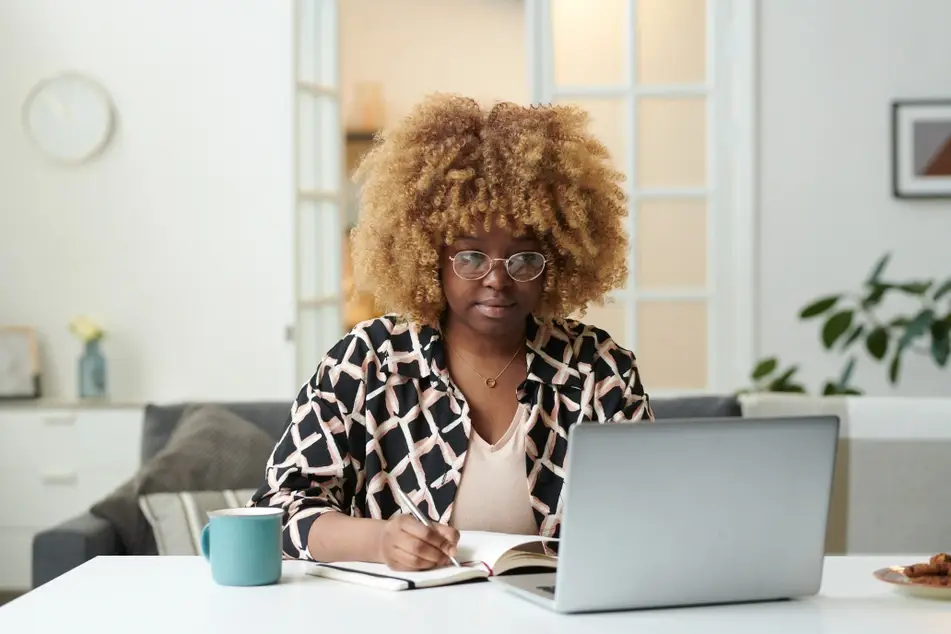 A Black woman with a blonde afro sits at a desk in her neat living room as she looks at an open laptop screen and writes in a notebook. There's a blue mug on the table next to her, and she is wearing a patterned blouse.