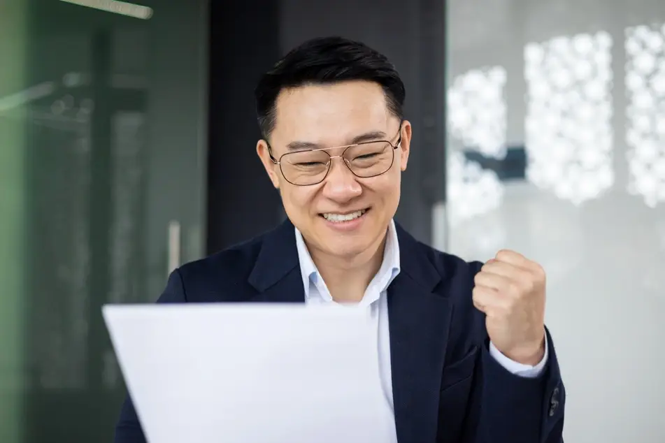 Older Asian man looks at job offer and pumps fist in the air in celebration in office.