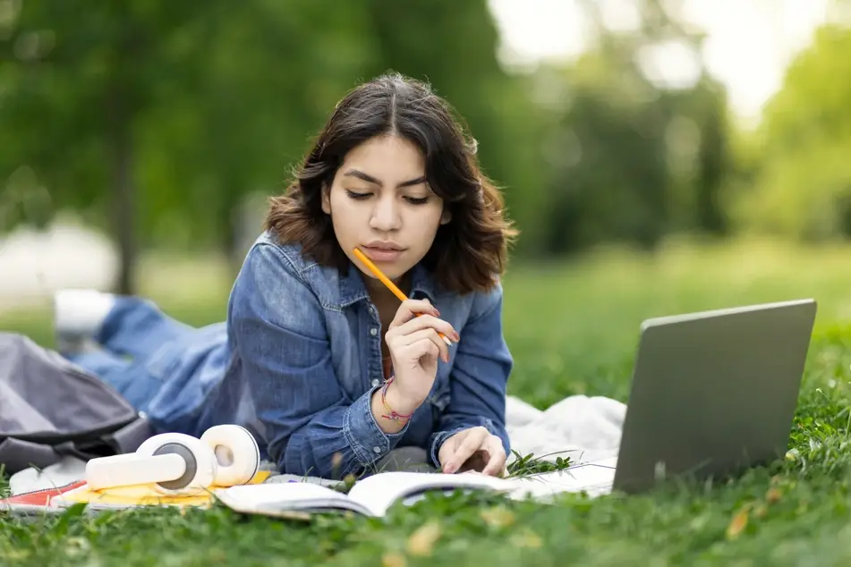 A photograph of a young woman sitting on grass while studying with open books and a laptop.