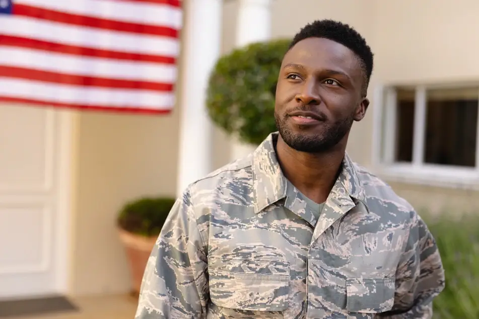 A photograph of a Black man in a camouflage uniform standing in front of a house, with a U.S. flag in the background.