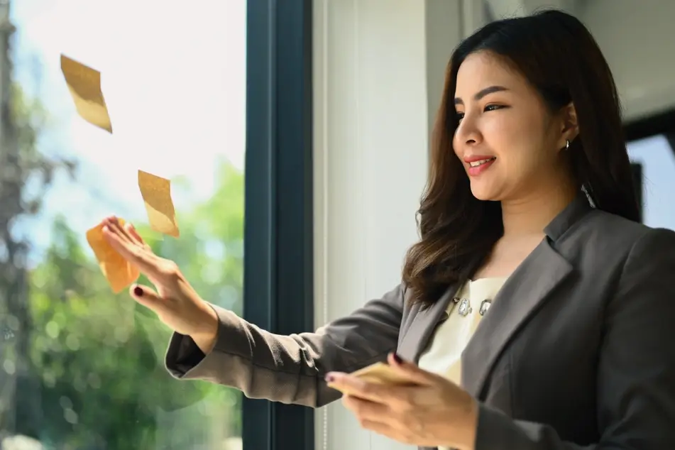 A woman wearing business clothing stands in front of a window in an office. She is attaching orange sticky notes to the glass.