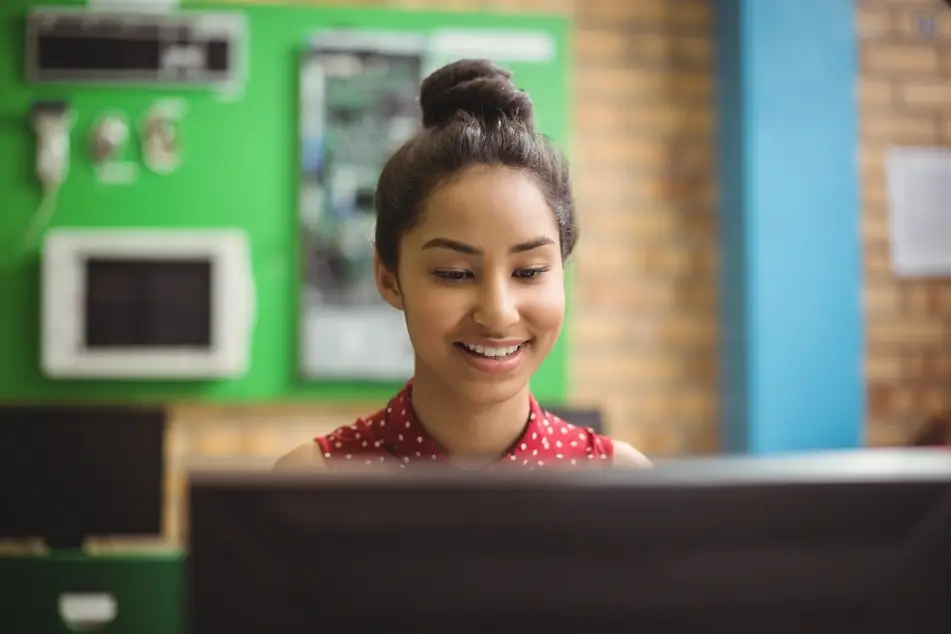 A young woman with dark hair sits at a computer screen in a technology office. She is smiling and wearing a red and white polka-dotted blouse.