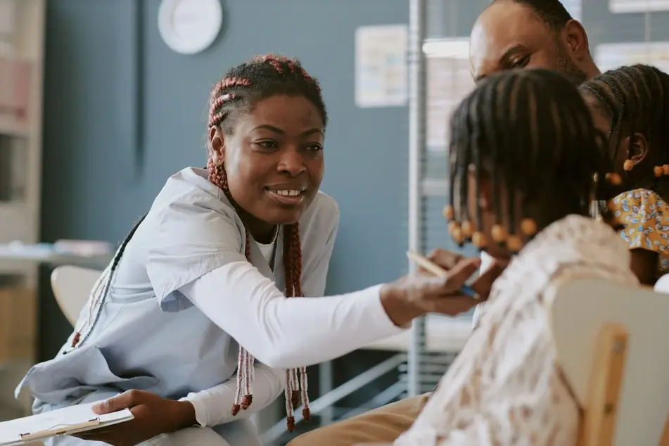 Black woman smiles while treating a small child in an office while wearing scrubs.