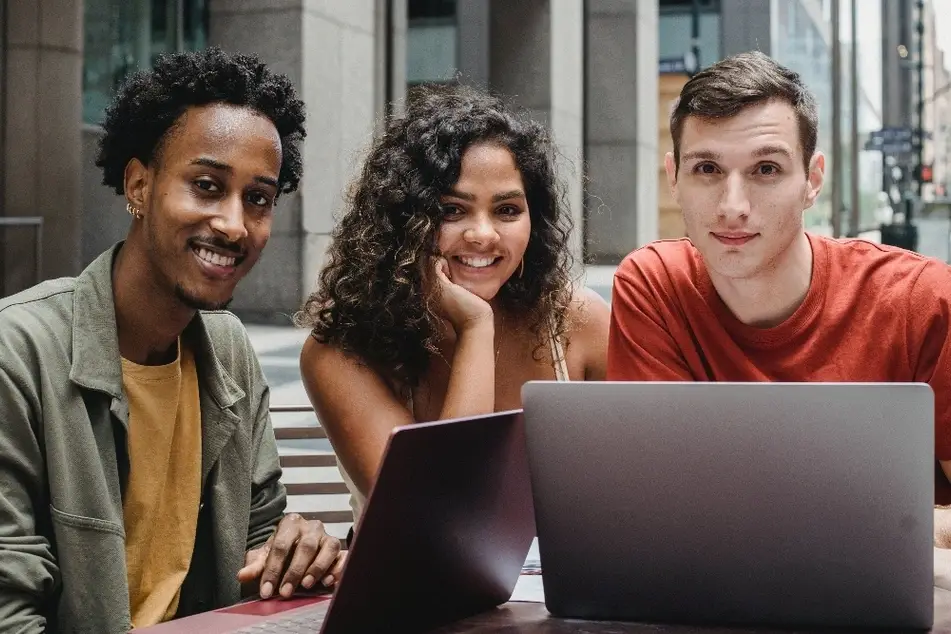 Tres jóvenes sentados frente a unas computadoras viendo a la cámara.