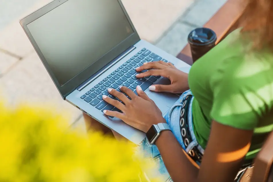 Black woman types on social media during her job search while sitting on a bench outside, wearing a bright green shirt and jeans.