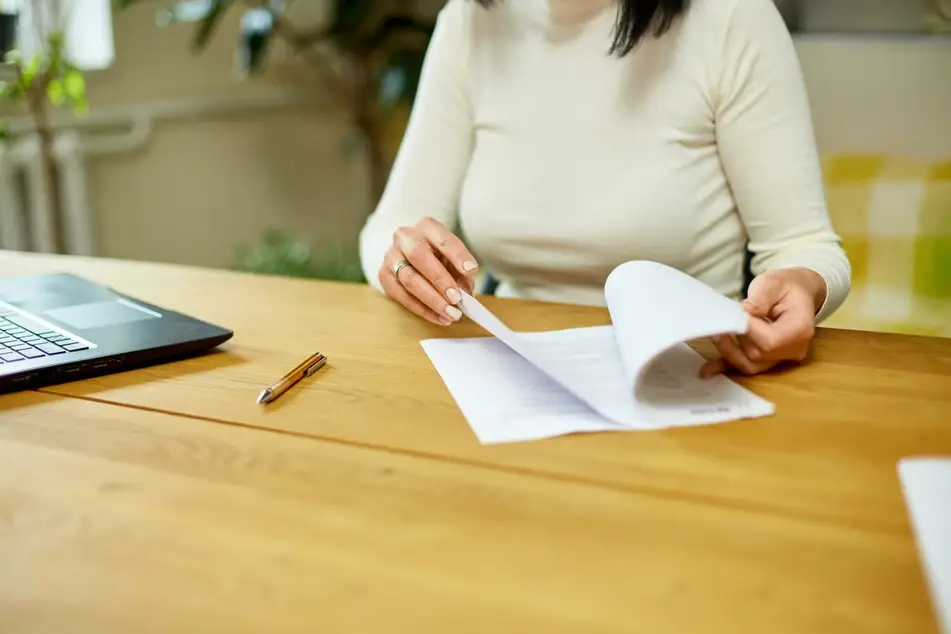 A woman in a long-sleeve, cream-colored shirt flips through a stack of papers on her wooden desk, for Idealist's blog post about understanding Form 990 to learn more about nonprofit compensation.