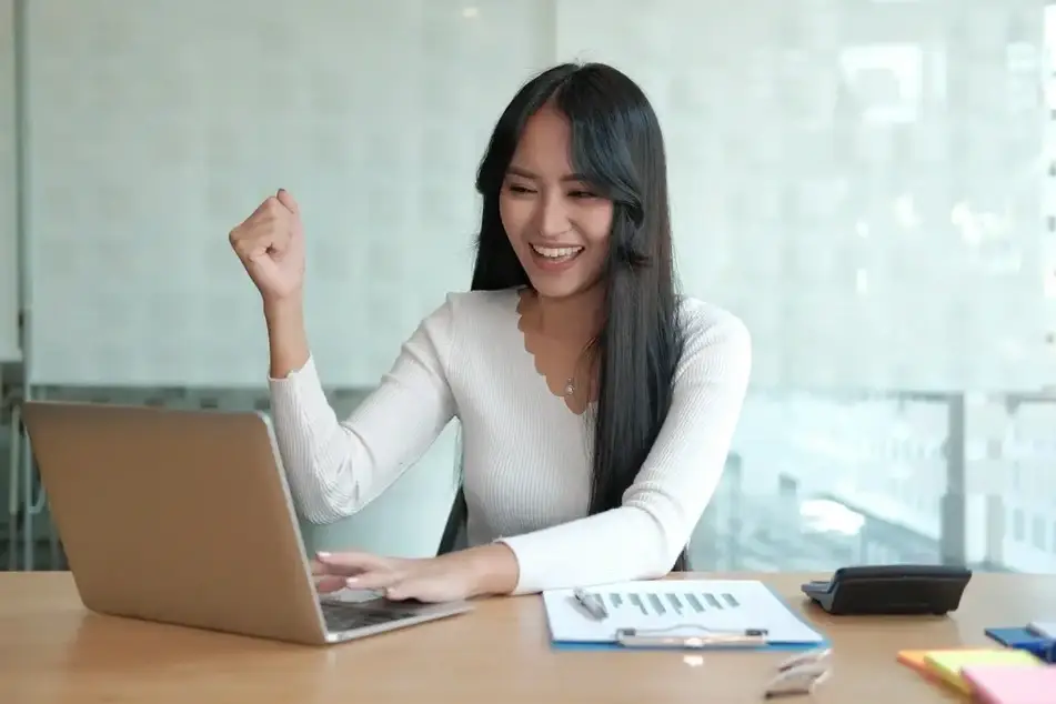 Foto de uma mulher de cabelo longo liso, com blusa branca de manga longa, sentada em frente de um notebook e uma prancheta com uma folha com gráficos e uma calculadora, com a mão fechada como se estivesse conseguido ou acertado na carreira