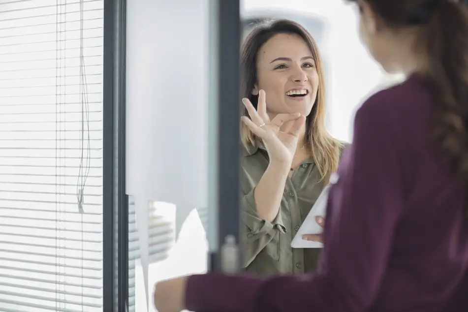 Two women talking at office. One white woman smiling and holding her hand up to the other, whose face is turned away.