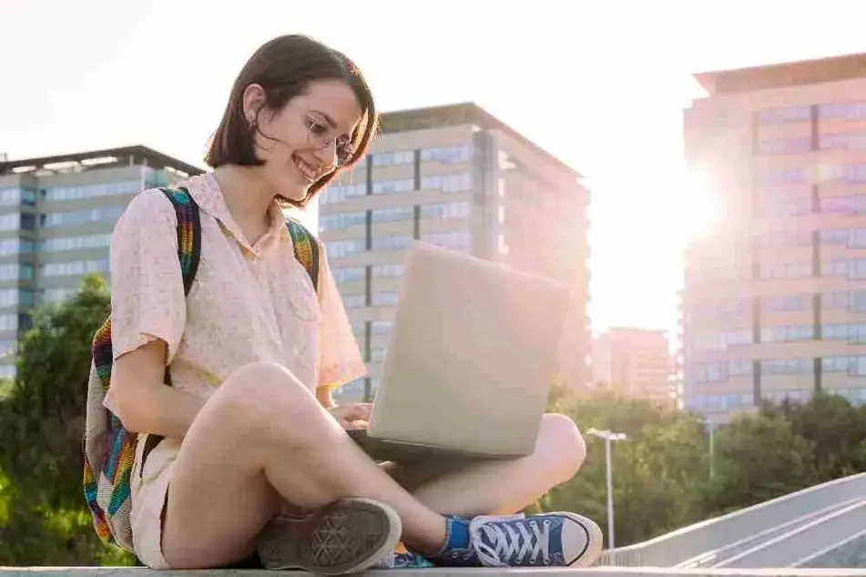 Una joven está sentada en el exterior trabajando sonriente en su computadora