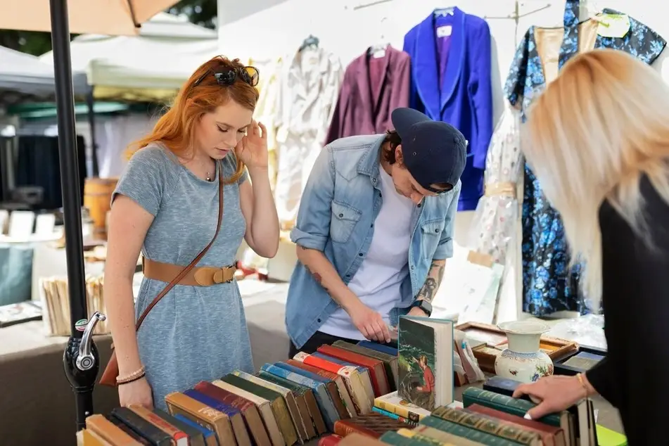 Imagem com 3 pessoas interagindo com livros que estão em um balcão. Ao fundo estão algumas pessoas de roupa.