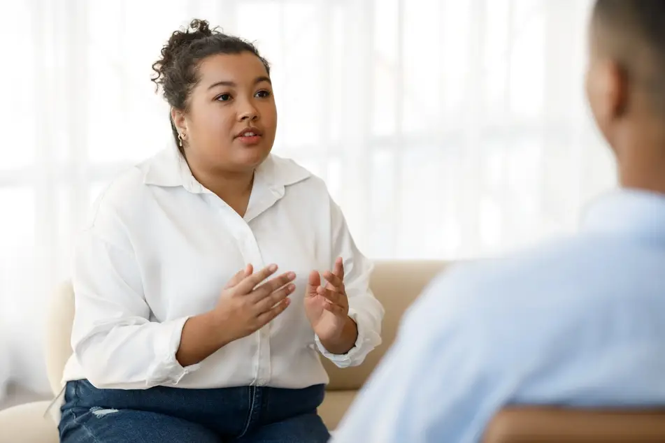 A young woman in white button down speaks to someone sitting across from her while sitting on a couch.