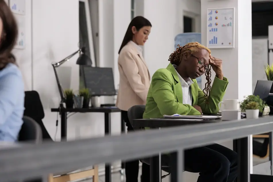 Black woman wearing a bright green blazer sits at desk in busy office, looking unhappy and unmotivated at work.