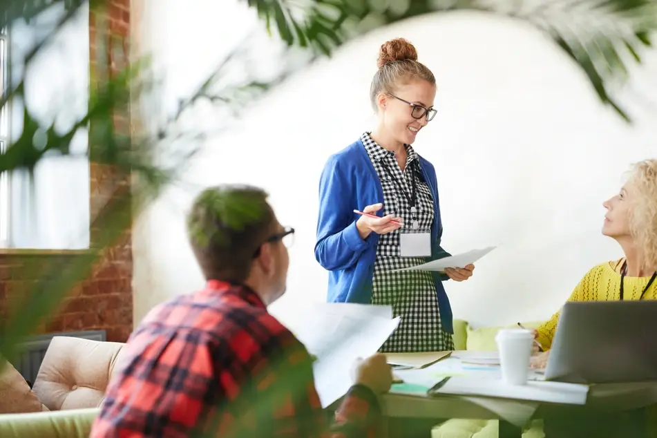 A photograph of several co-workers in an office, as their manager stands around them smiling and talking about work.