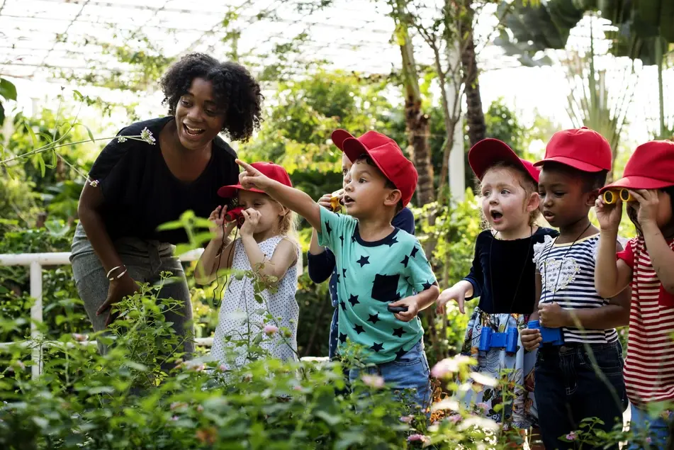 A photograph of a Black teacher working in education takes her Kindergarten students to a plant nursery to learn about ecology.