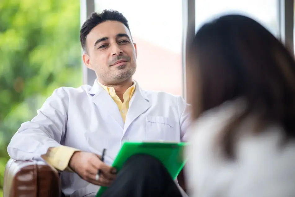 A photograph of a male doctor sitting in a chair as he talks to a woman sitting across from him.