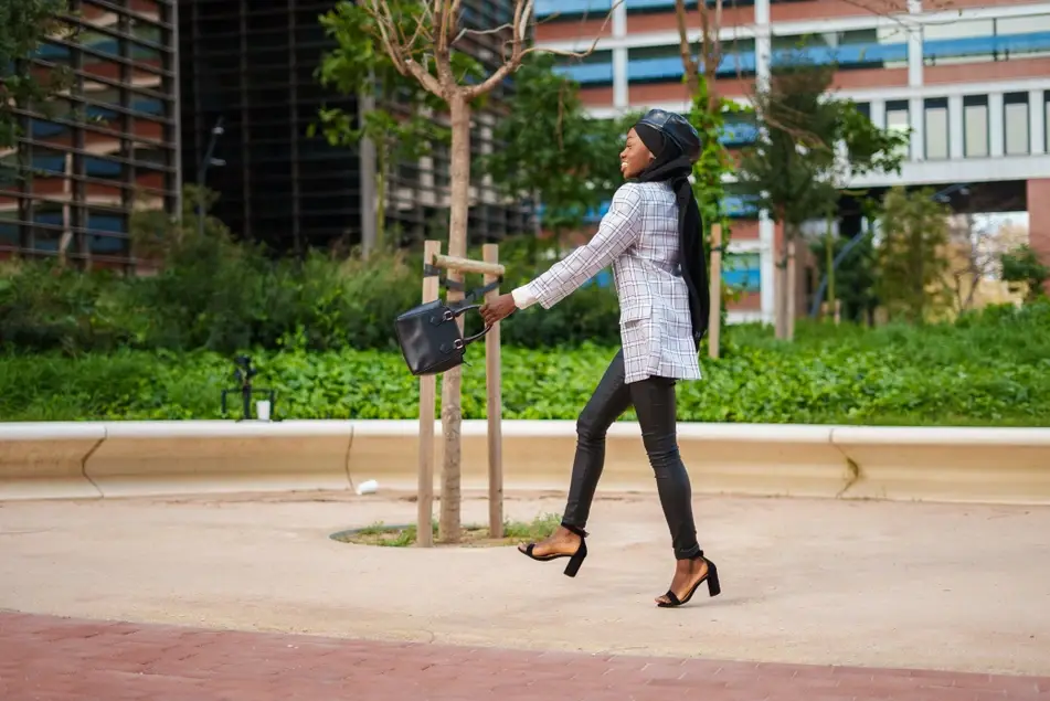 Black woman wearing a head scarf, hat, heels, and a blazer happily walks outside swinging a purse.