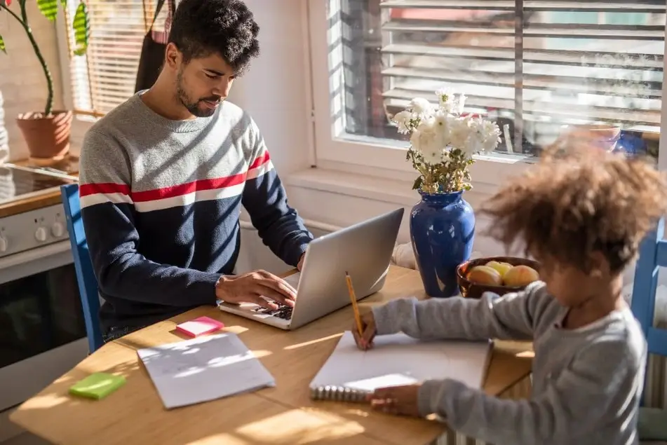 Un hombre joven trabaja desde casa con su computadora mientras cuida de una niña.