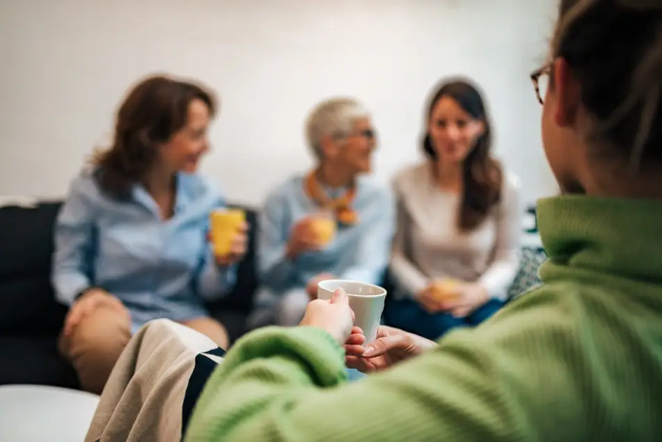 A group of four women are sitting on a couch in a room talking with coffee cups in their hands.