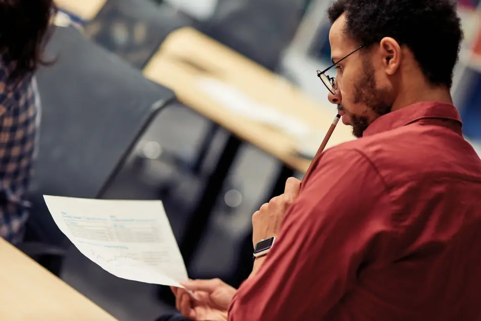 Black man wearing a red shirt and apple watch sits at home looking at grad school student loan paperwork, holding a pencil to his lips.