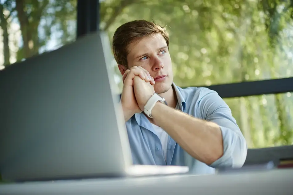 White man sits at office desk thinking, green trees in the background.