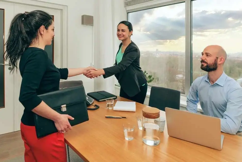 Dos mujeres se dan un apretón de manos mientras un hombre las observa sonriente durante una entrevista de trabajo.