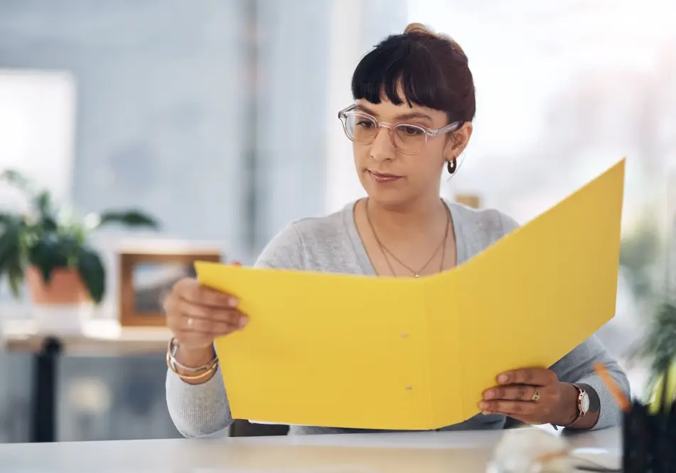 Woman wearing glasses holds a yellow file in an office environment.