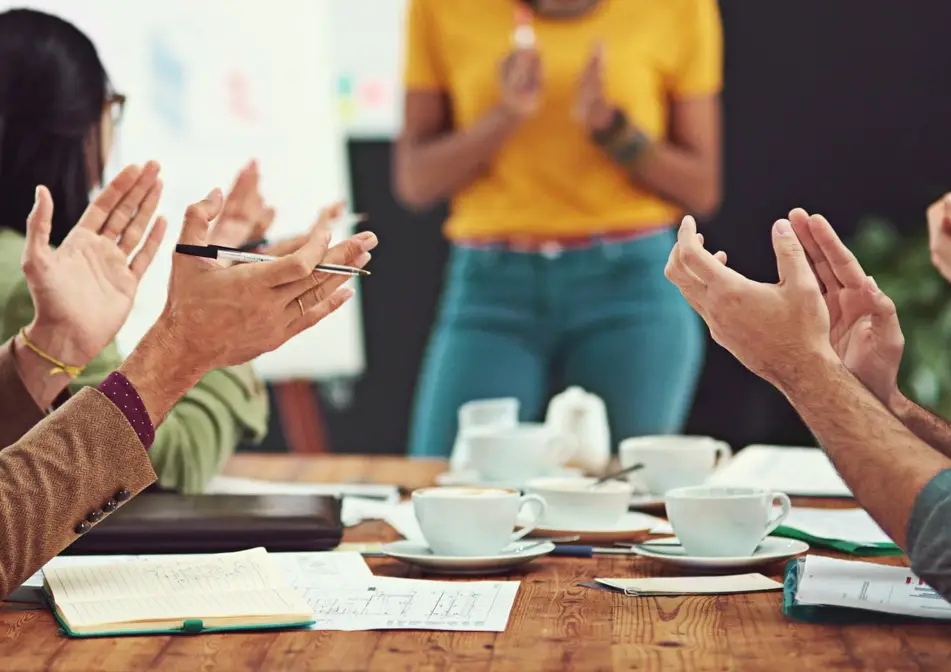 Group of co-workers applauding someone during a meeting, coffee cups strewn across the table.